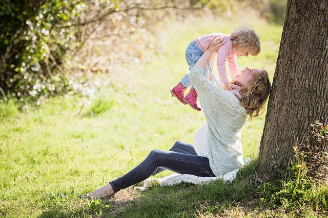 Happy pregnant mother lifting up her daughter while sitting and leaning against a tree